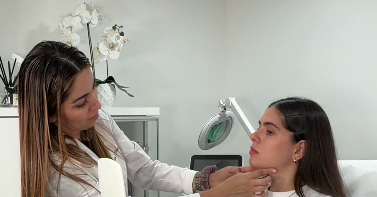 A clinician in a white coat gently checks a client's jawline and cheek in a bright ROW 89 treatment room with white orchids and a magnifying lamp