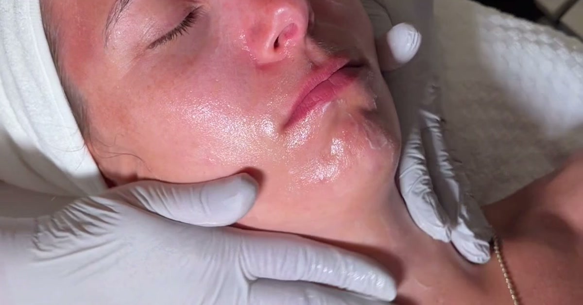 An esthetician's gloved hands massage a glossy facial product along a client's jaw and cheek during a facial at ROW 89, the client's hair wrapped in a white towel