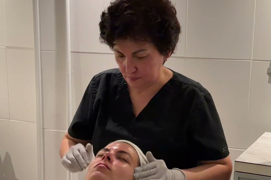 An esthetician in black scrubs and gray gloves gives a client a facial in a white-tiled treatment room at ROW 89.